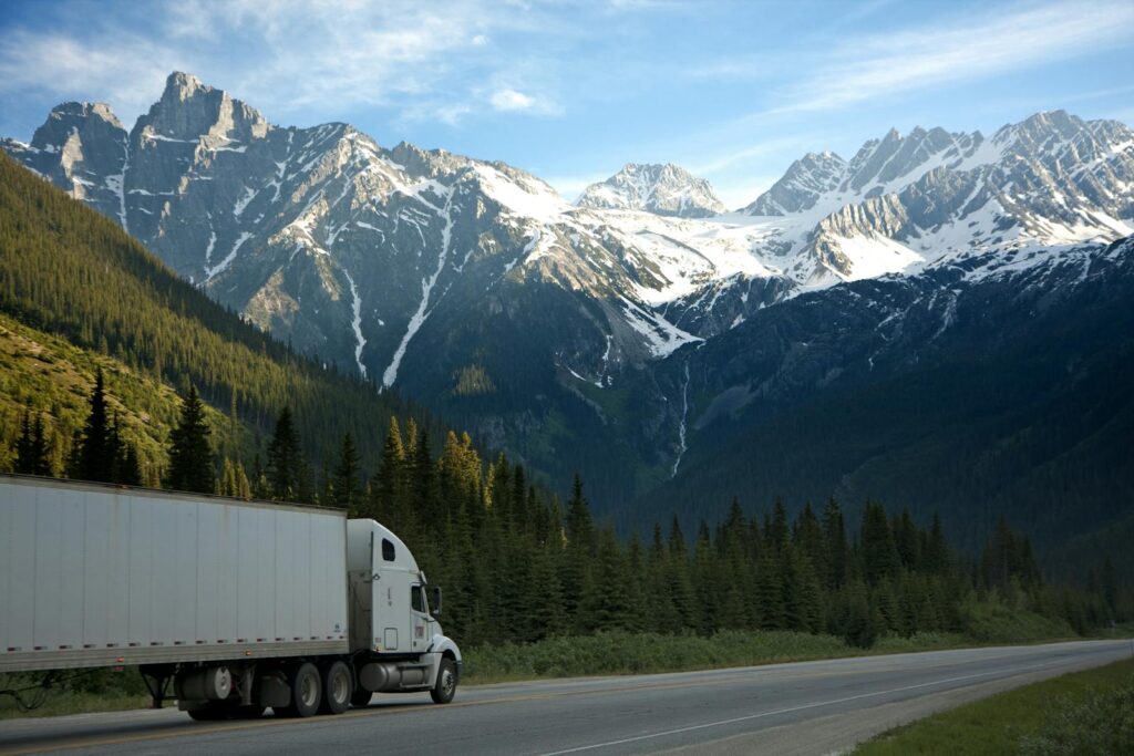 A semi-truck travels along a highway with snow-capped mountains in the background.